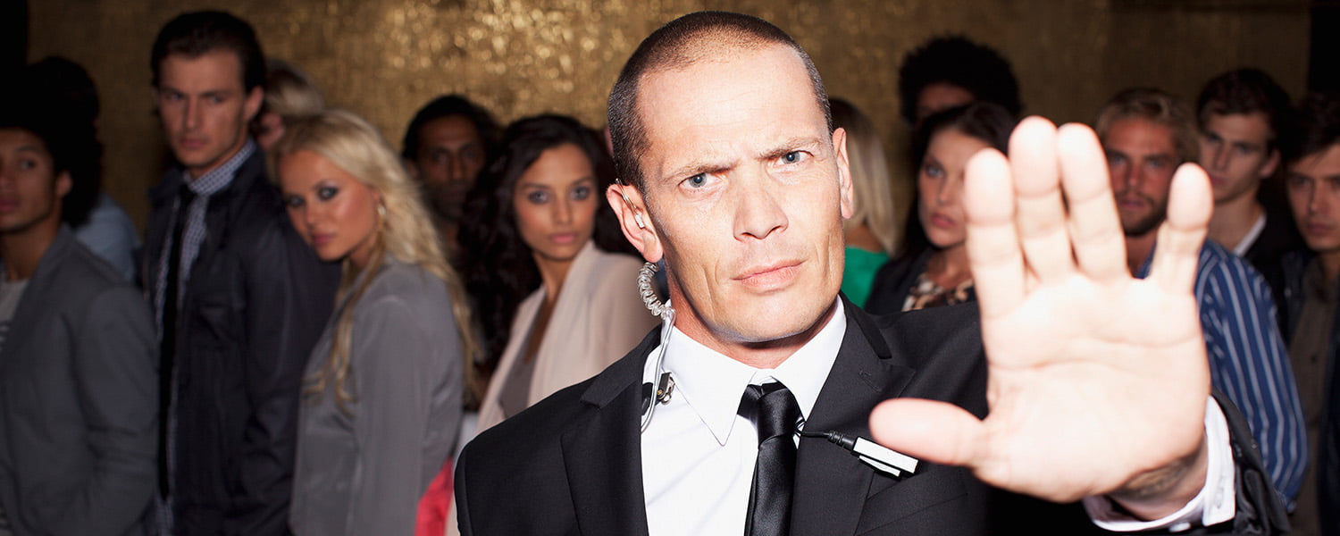 A security guard dressed in a suit holding up his hand to block entrance with partygoers in the background